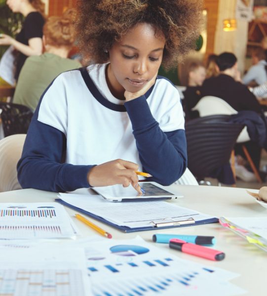 young-african-woman-entrepreneur-with-serious-concentrated-expression-sitting-coworking-cafe-with-touch-pad-pc-papers-analyzing-financial-information-tablet-resting-elbow-table
